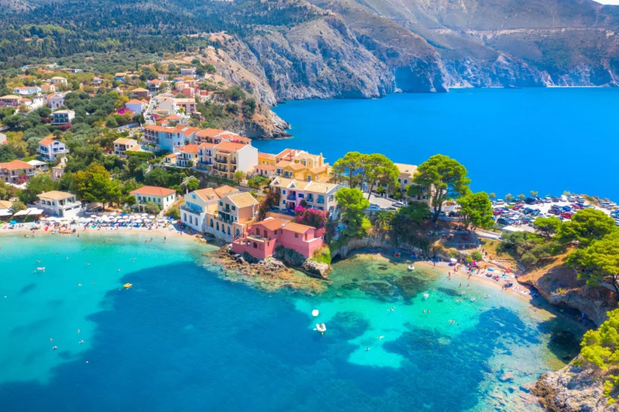 Mediterranean village beach scene with turquoise waters, sunbathers, colorful buildings, and cliffs.