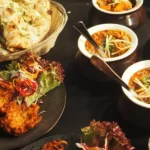 Vibrant Indian cuisine display: naan, curry bowls, onion bhaji, and mint chutney on dark background.
