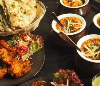 Vibrant Indian cuisine display: naan, curry bowls, onion bhaji, and mint chutney on dark background.
