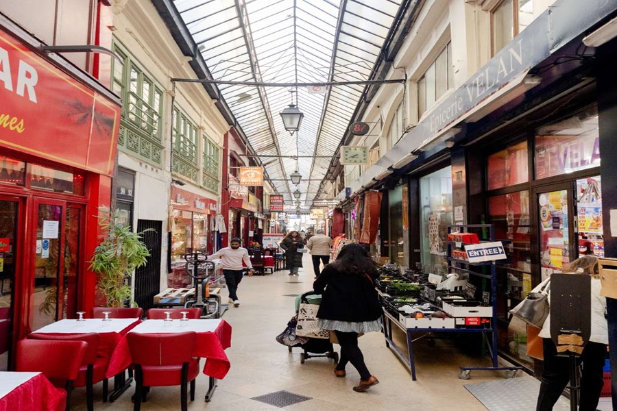 Vibrant market arcade with shops, eateries, glass ceiling, ornate railings, and diverse patrons.