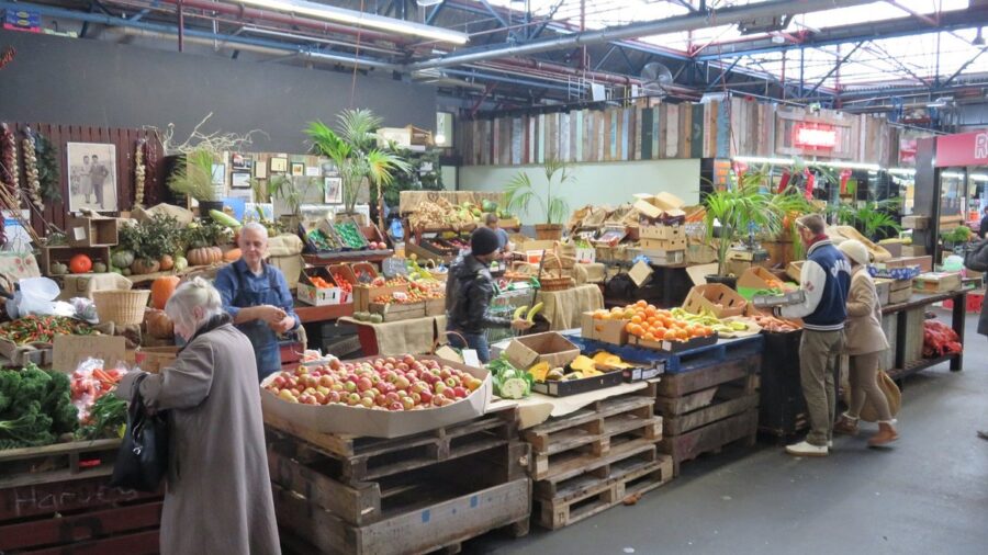 Indoor farmers market produce stall with fresh vegetables, fruits, vendors, and shoppers under rustic industrial lighting.