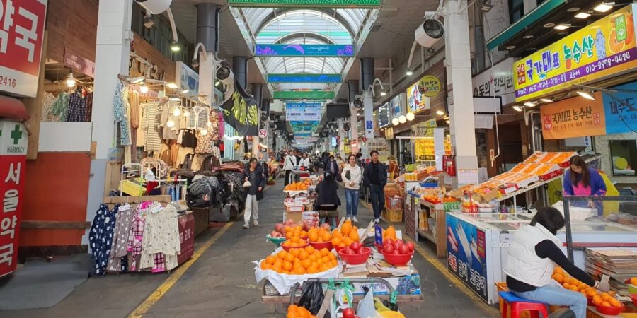 Busy Seogwipo Maeil Olle Market in Korea with indoor fruit stands and clothing stalls.