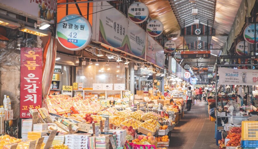 Busy Korean indoor market with colorful fruit stalls, produce vendors, and shoppers under bright hanging signs.