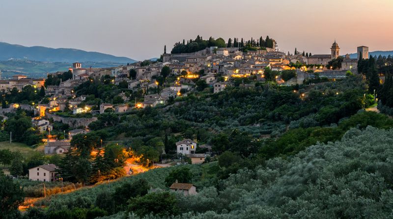 Italian hillside town at dusk with stone buildings, cypress trees, and twilight sky.