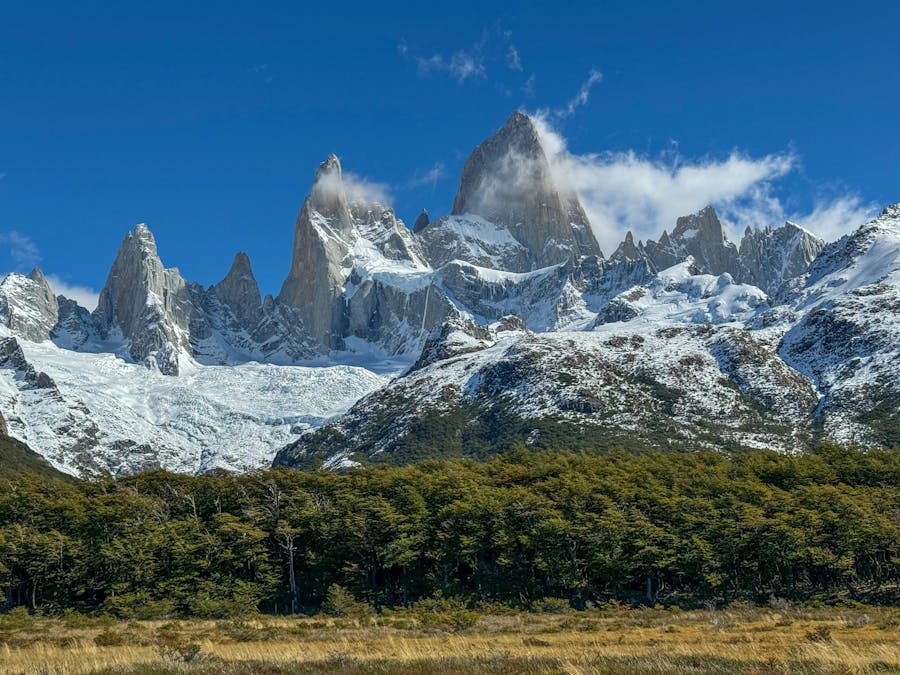 Snowy jagged mountain range with towering peak above forest and autumn meadow landscape.