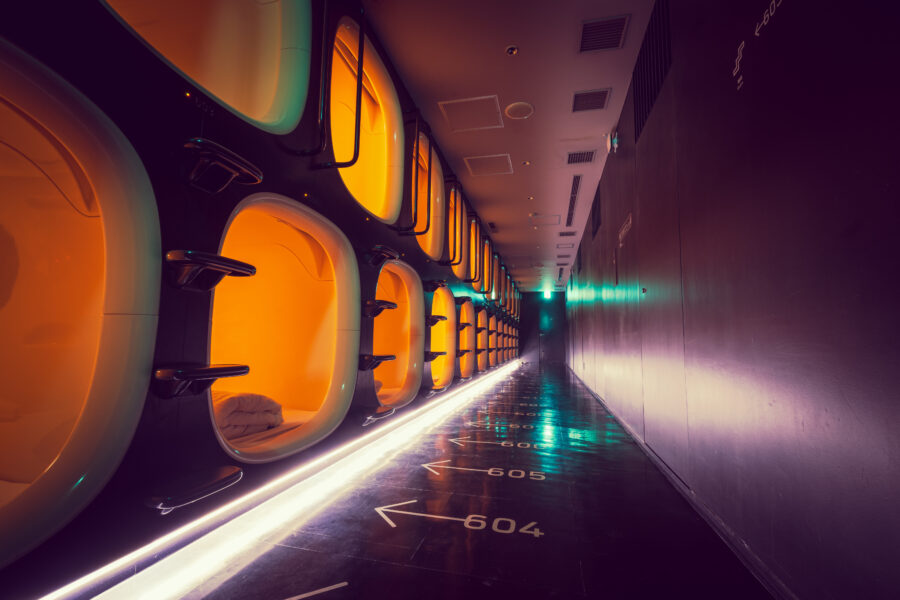Dimly lit futuristic capsule hotel hallway with stacked pods on the left, glowing with warm orange light. The floor has numbered arrows and a sleek, modern design.
