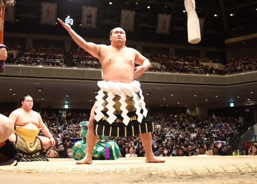 Sumo wrestler performing a pre-match ritual on a dohyo, with a large audience in an indoor arena. The mood is intense and focused.