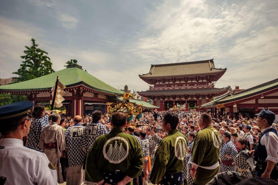 Japanese festival at temple with mikoshi, traditional attire, crowded scene, cultural celebration.