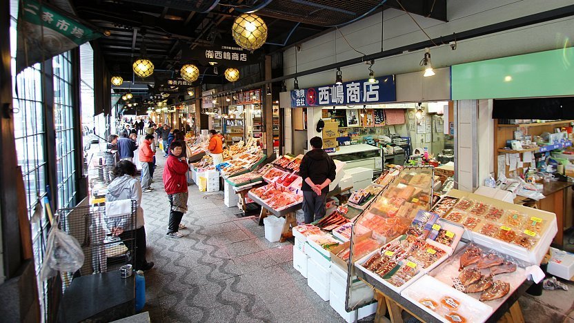 Busy indoor Japanese seafood market with fresh fish stalls, vendors, and shoppers along a covered walkway.