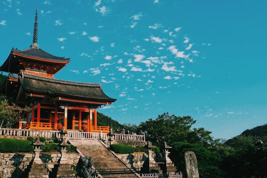 Traditional Japanese temple against blue sky with clouds, surrounded by lush greenery and stone lanterns.
