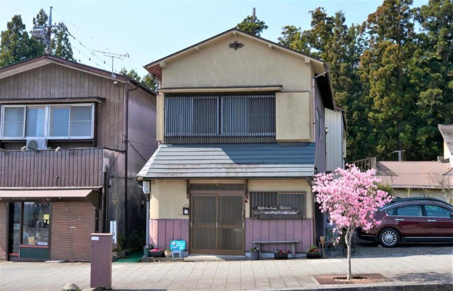 Traditional Japanese guesthouse street view in spring with blooming tree, wooden facade, and parked car.