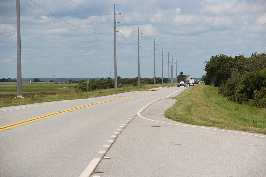 Vehicles drive along the Jekyll Island Causeway, with power lines visible alongside the road.