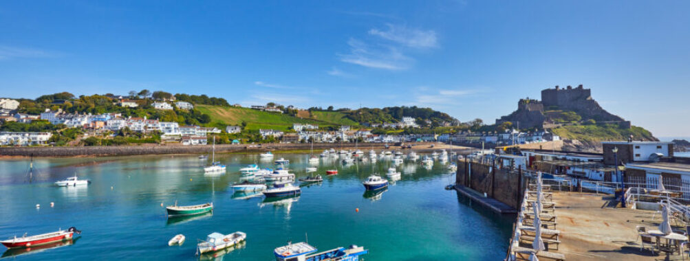 Gorey Harbour in Jersey Channel Islands, featuring fishing and pleasure boats, with Gorey Castle and a clear blue sky in the background.