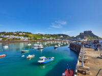 Gorey Harbour in Jersey Channel Islands, featuring fishing and pleasure boats, with Gorey Castle and a clear blue sky in the background.
