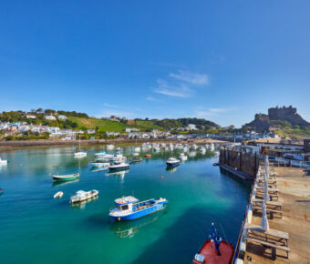 Gorey Harbour in Jersey Channel Islands, featuring fishing and pleasure boats, with Gorey Castle and a clear blue sky in the background.