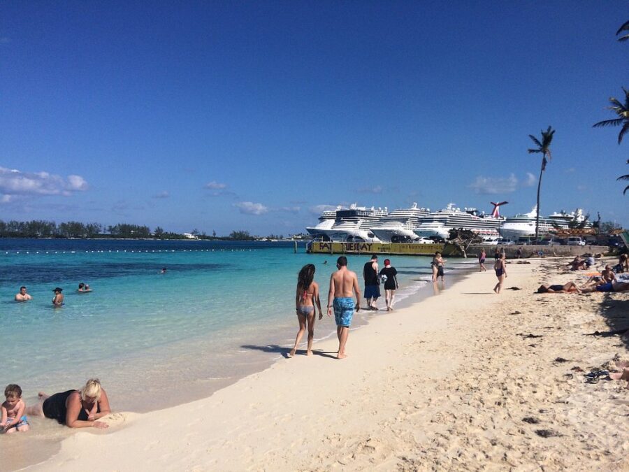 Junkanoo Beach scene with people walking on soft sand and swimming in clear blue water. Several large cruise ships are docked in the background.