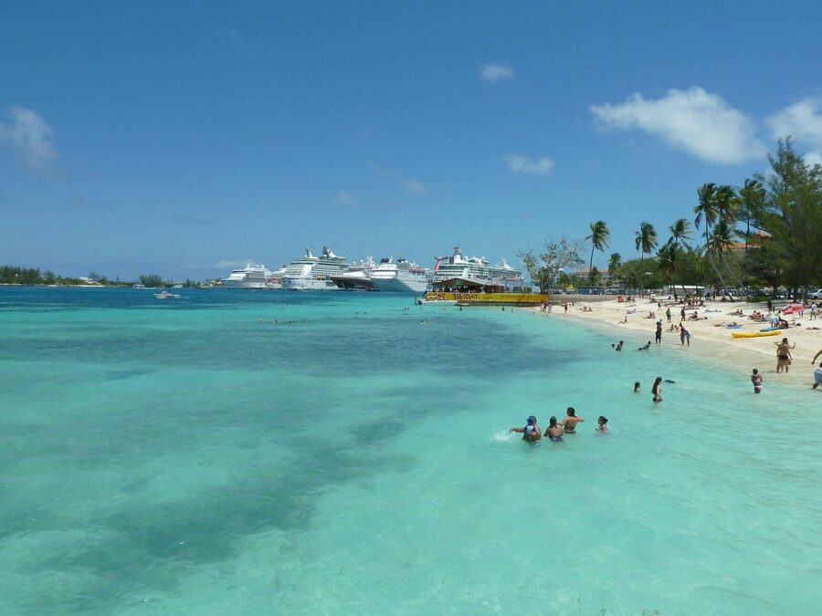 Clear turquoise water and Junkanoo Beach with scattered palm trees, sunbathers, and swimmers; cruise ships docked in the distance under a bright blue sky.