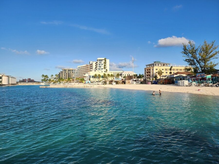 Beachfront of Junkanoo Beach with clear blue water, hotels, and a lively shore. Two people swim, while buildings and palm trees line the sunny, serene coastline.
