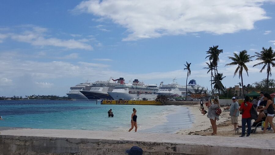 Beach scene with people enjoying the sand and sea. Cruise ships are docked in the background. Palm trees line the shore, under a partly cloudy sky.