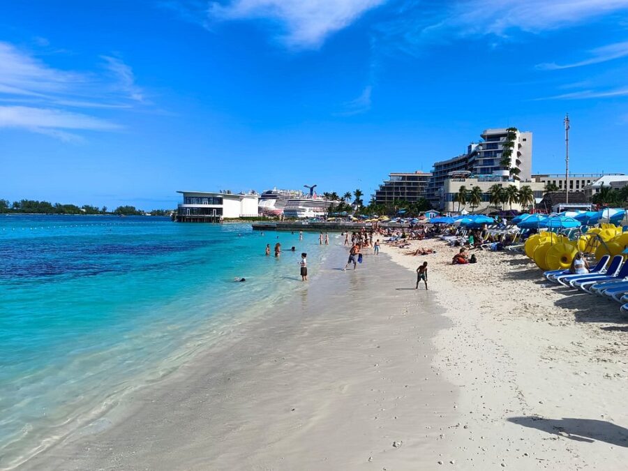 Junkanoo Beach with turquoise waters, people swimming and lounging under blue umbrellas. Cruise ships and modern buildings line the horizon.
