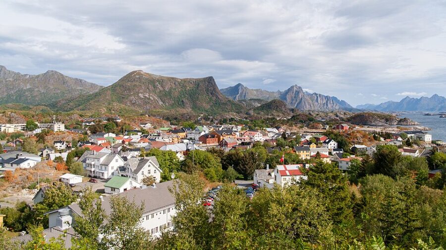 Scenic view of Kabelvåg, Norway with colorful houses surrounded by lush greenery, set against rugged mountains and a cloudy sky, conveying tranquility.