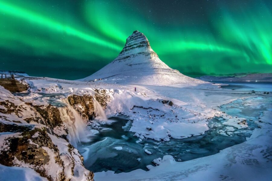 Kirkjufell mountain in Iceland under aurora borealis with snow, waterfall, and icy river.