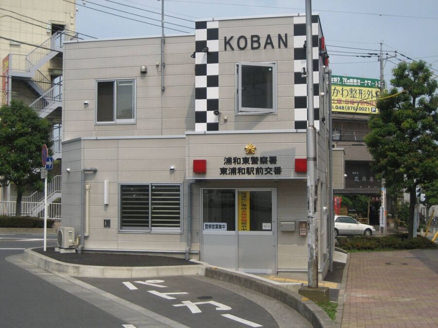 Modern Japanese Koban police station in urban setting with pedestrian path and greenery.