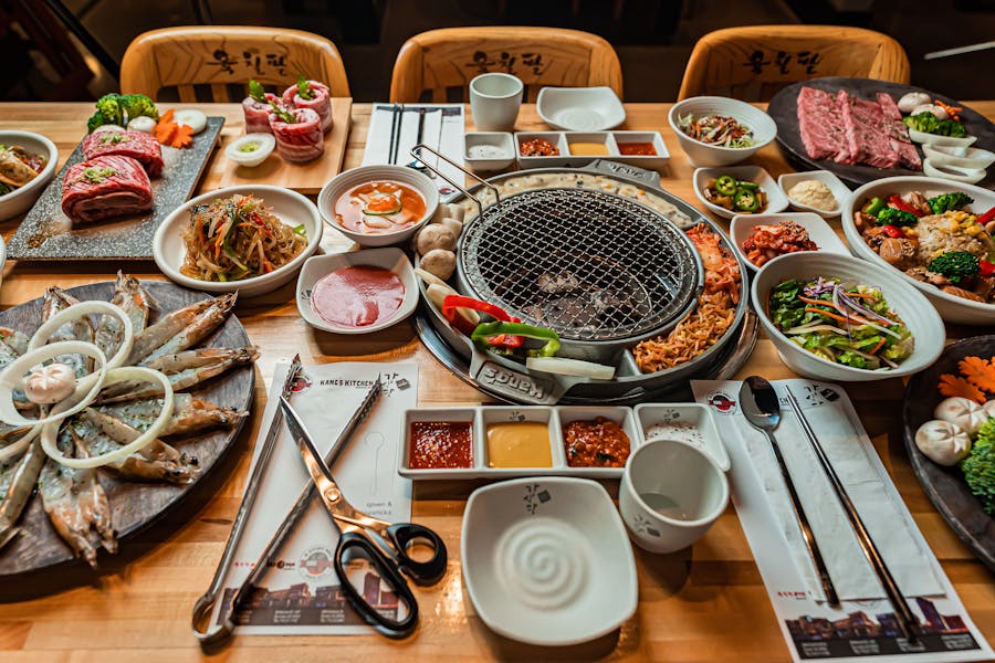 Korean BBQ grill setup with meats, vegetables, banchan, and dipping sauces on a wooden table.