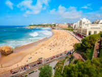 Sunny day on La Grande Plage Beach, Biarritz, France, lined with sunbathers and swimmers. A coastal cityscape with green cliffs and a distant lighthouse under blue skies.