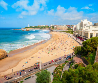 Sunny day on La Grande Plage Beach, Biarritz, France, lined with sunbathers and swimmers. A coastal cityscape with green cliffs and a distant lighthouse under blue skies.