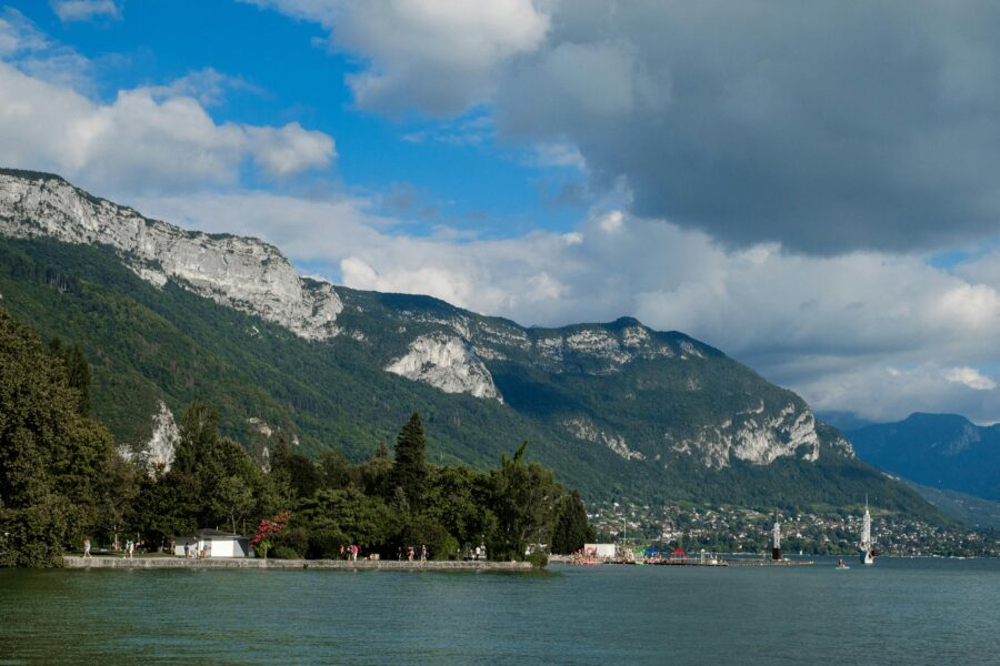 Lake Annecy shoreline with Alps mountains, promenade, lighthouse, and sailboat.