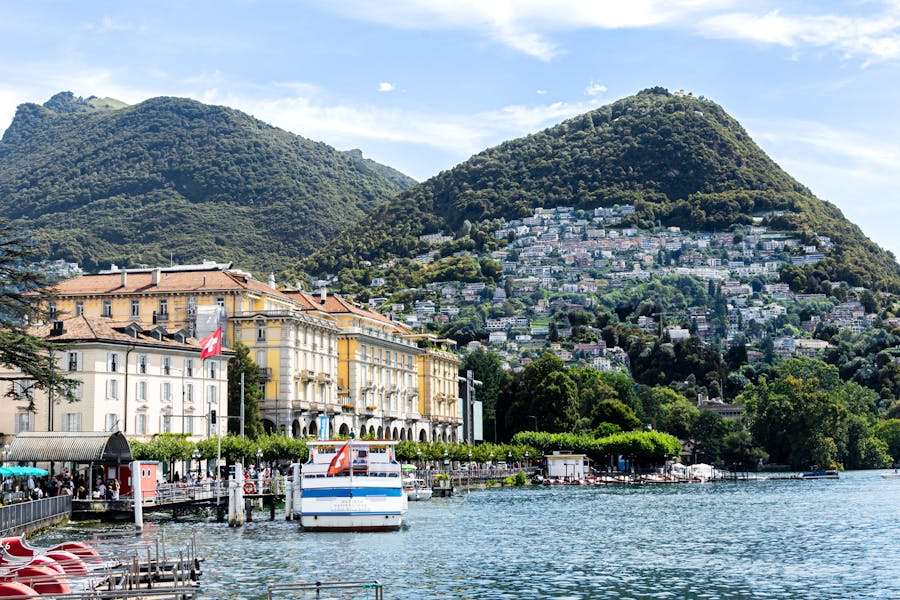 Lugano lakefront with ferry, pedal boats, waterfront promenade, hillside villas, Swiss flag.
