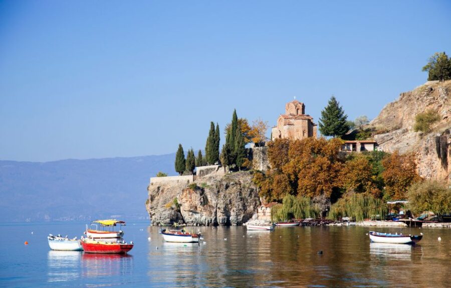 Tranquil Lake Ohrid with boats, cliffs, autumn trees, and historic cliffside church.