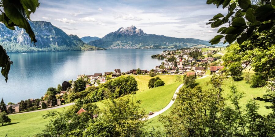 Tranquil lake with lush hills, village, and majestic mountains reflecting sky and clouds.