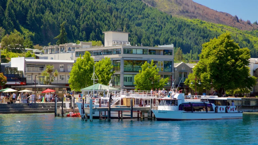 Queenstown lakefront boat dock with tourist cruise ship, waterfront cafes, modern buildings and forested mountains.