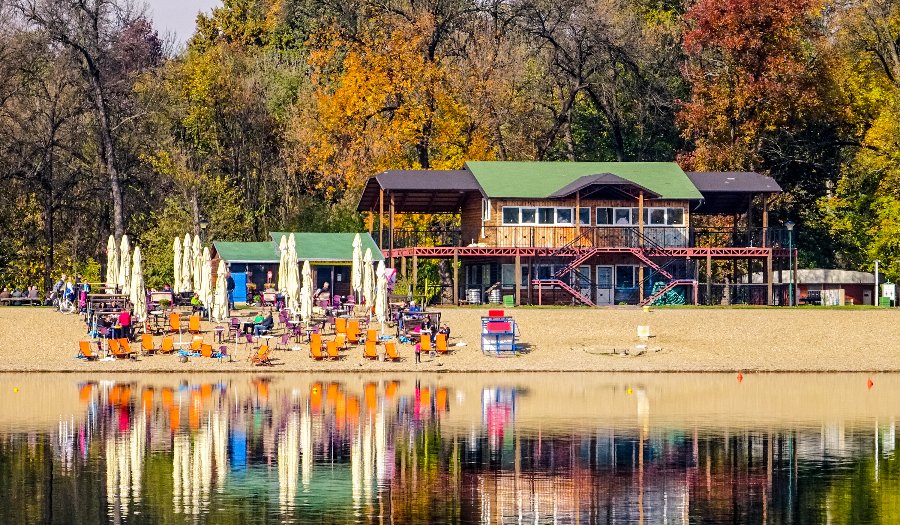 Serene lakeside autumn scene with beach, lounge chairs, colorful trees, and rustic building.