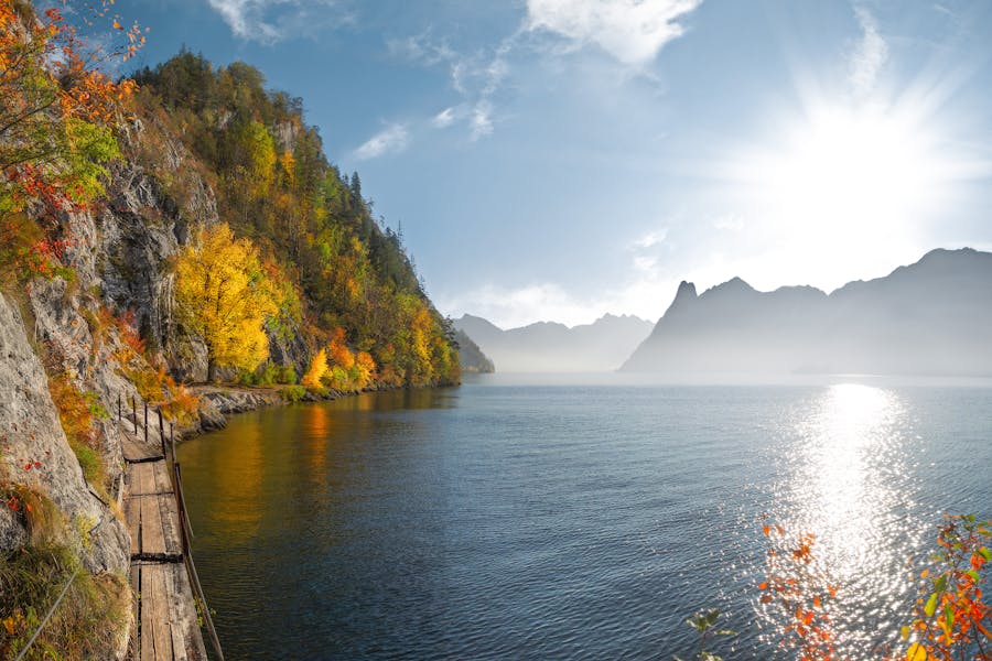 Autumn lakeside boardwalk along cliff with sunlit water and distant mountains.
