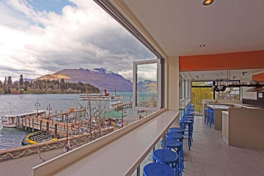 Modern lakeside café interior with bar seating overlooking mountain lake, boats, pier, and scenic harbor.