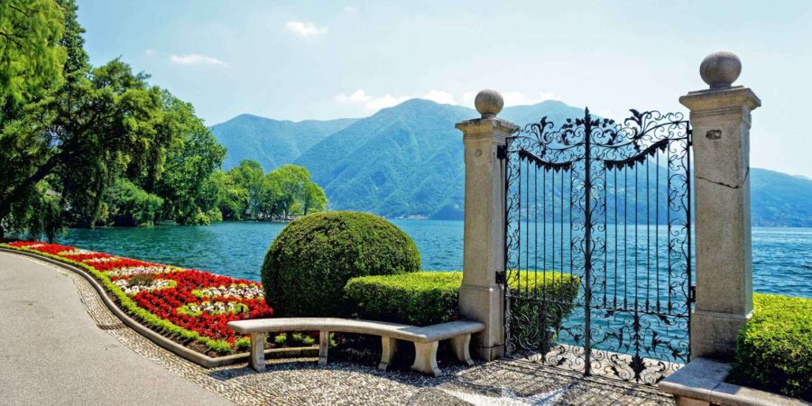 Parco Ciani lakeside garden gate opening to lake and mountains, promenade with bench and flowers.