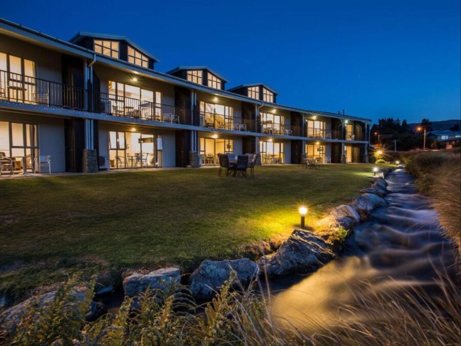Lakeside motel lodge at dusk with illuminated rooms, garden lawn, outdoor seating, and stream.