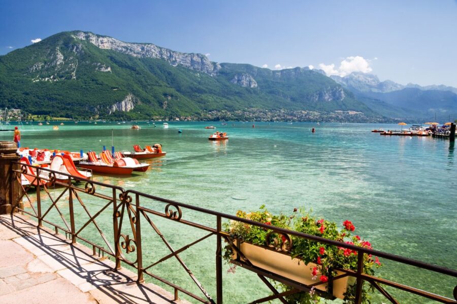 Lakeside promenade with turquoise lake, red paddleboats, mountain backdrop and flowering railing planter.
