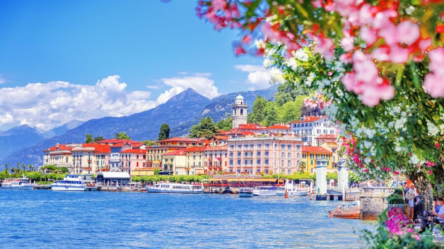Lake Como lakeside town with colorful waterfront buildings, moored boats, bell tower and mountains.