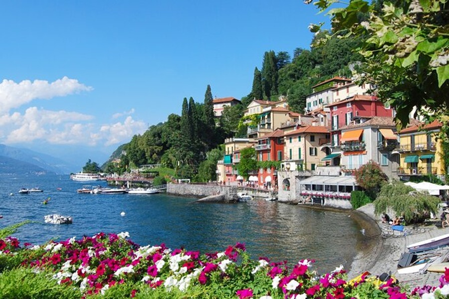 Lake Como waterfront village promenade with colorful buildings, boats, cypress trees, and flowers.