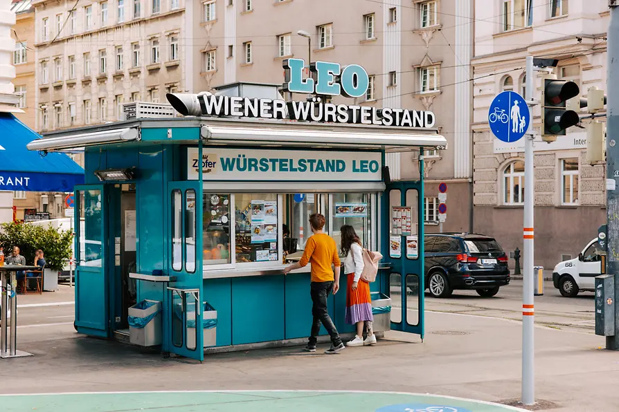 Teal Leo Würstelstand kiosk serving street food to customers on a Vienna city sidewalk.