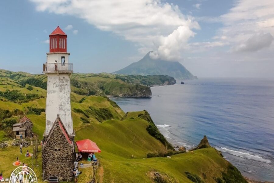 White lighthouse with red lantern on coastal cliff, green hills, rocky shore, distant mountain.