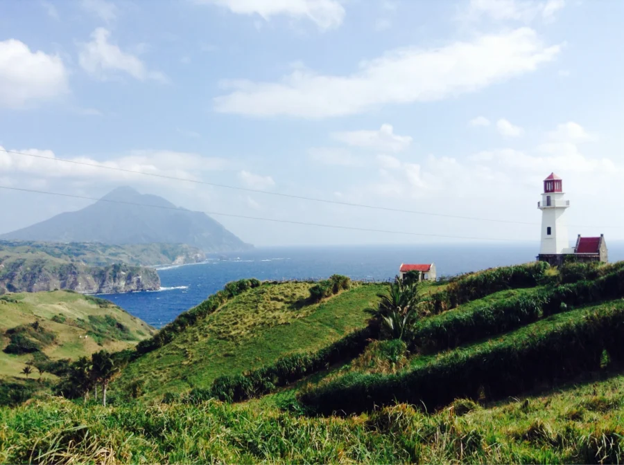 Tayid Lighthouse on grassy coastal cliffs overlooking blue ocean and distant island.
