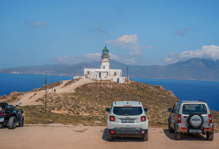 Lighthouse on rocky Aegean coast, Mykonos, with parked vehicles and tourists exploring.