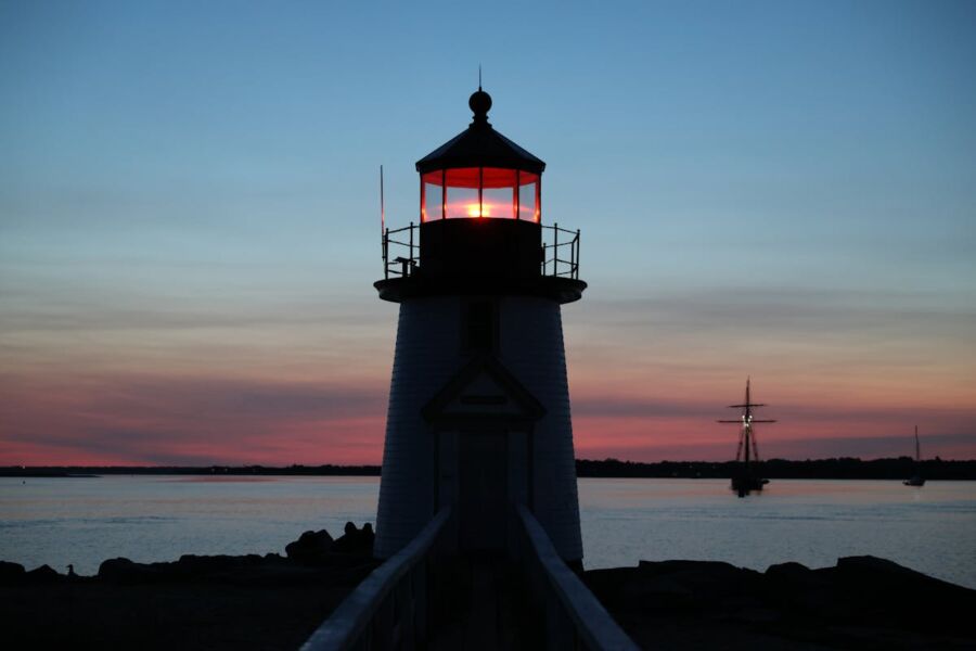 Nantucket Twilight lighthouse with glowing light, rugged rocks, sailboat, and colorful evening sky.