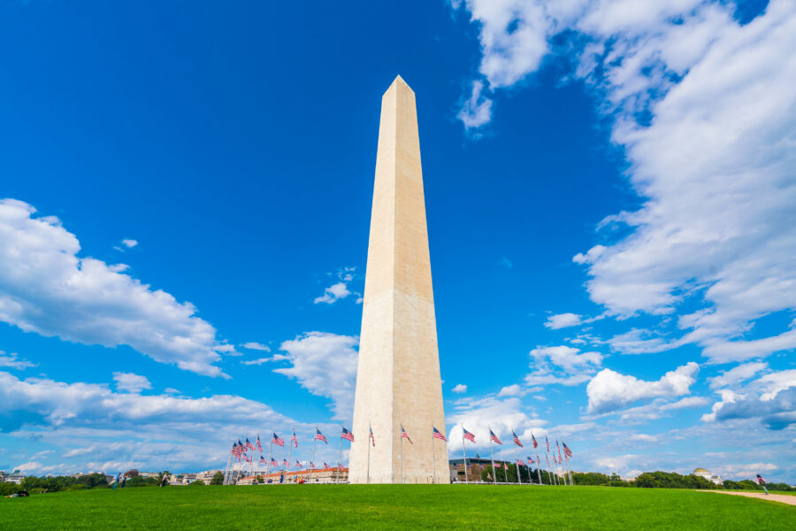 The Washington Monument stands tall against a vivid blue sky with scattered clouds, surrounded by a circle of waving American flags on a lush green lawn.