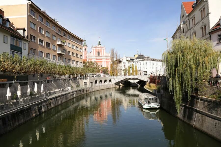 The Ljubljanica River flows through a city, surrounded by trees and buildings, showcasing urban life.